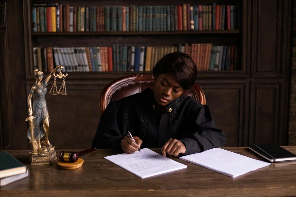 A judge in robes writing on a document at a desk in an office library with law books.