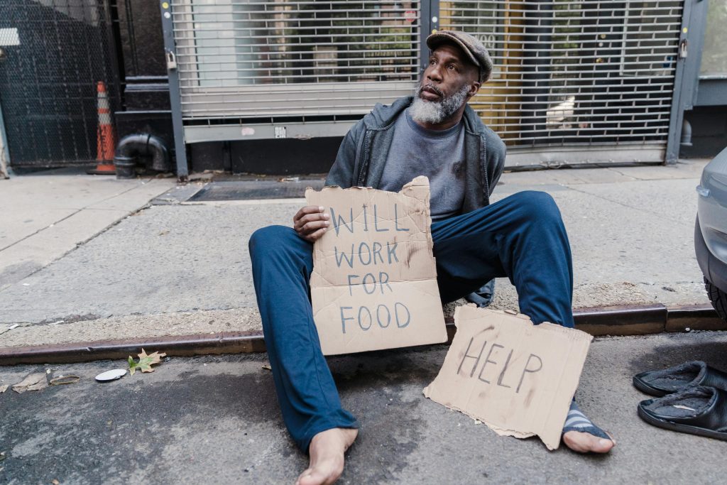 Homeless man sitting on sidewalk holding signs for help and work on a city street.